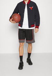 Schwarze Collegejacke mit roten Streifen und Logo, weißes T-Shirt, schwarze Shorts mit grauen und roten Akzenten, schwarze Turnschuhe, hält einen Basketball.