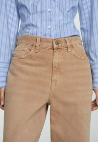 Jeans di cotone color sabbia con taglio dritto, dotati di passanti per cintura, tasche frontali e chiusura clasica con bottone. Abbinati a una camicia a righe blu chiaro.