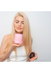 Różowy cylindryczny pojemnik z logo i błyszczącym wykończeniem, trzymany w jednej ręce, obok okrągłego czarnego kompaktu z metalicznymi akcentami.