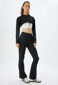 Schwarzer, verkürzter Hoodie über einem weißen Crop-Top, kombiniert mit schwarzen, ausgestellt geschnittenen Hosen und grauen Sneakers mit weißen Streifen. Glattes Material und taillierte Passform.
