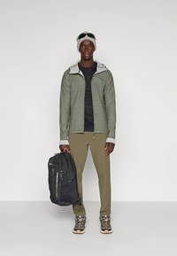 Moški nosi sivo kapo, sončna očala, zeleno zip-up jakno, črno majico, bež hlače in pohodne čevlje ter v eni roki drži črno nahrbtnik.