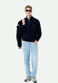 Mornarska pulover s zadrgo, v kombinaciji s svetlo modrimi džins hlačami in športnimi sivo-snežnimi čevlji. Črna varsity jakna je obešena čez en ramenski.