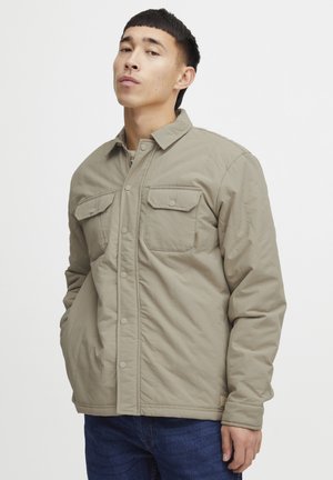 Khaki quilted jacket with button-up front, two chest pockets, long sleeves, and a straight cut; minimalistic design and textured fabric.