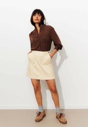 Chemise à boutons rayée brune, jupe en velours côtelé beige avec des poches avant, chaussettes grises et chaussures brunes avec lacets. Design simple et moderne.