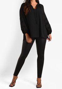 Svart tunika med prickar och V-ringning samt knytdetalj, med genomskinliga ballongsärmar. Matchas med glänsande svarta leggings och remsandaler.