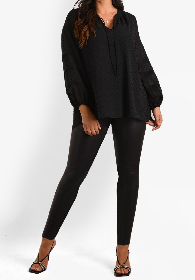 Svart tunika med prickar och V-ringning samt knytdetalj, med genomskinliga ballongsärmar. Matchas med glänsande svarta leggings och remsandaler.
