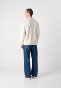 Kremfarget strikkegenser med rund hals, kombinert med løstsittende, midtblå denimjeans og hvite sko. Teksturert stoffdetalj synlig.