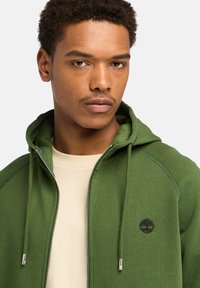 Grüne Zip-Hoodie mit weicher Textur, mit einem Logo auf der Brust und Kapuze mit Kordelzug. Darüber getragen ein beiges Shirt.