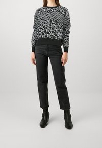 Mujer con un top de manga larga de patrón en blanco y negro, vaqueros negros de cintura alta y botines negros, de pie frente a un fondo liso.