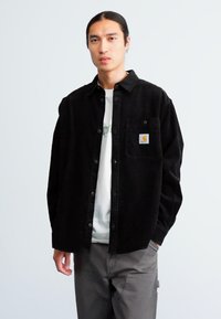 Chemise en velours côtelé noir avec une poche et un logo, design à boutons, portée par-dessus une chemise claire, associée à un pantalon gris.