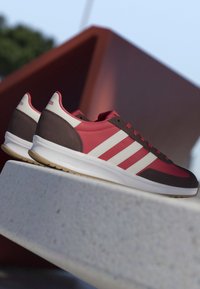 Rote und braune Sportschuhe mit weißen Streifen, die auf einer Betonfläche vor einem verschwommenen Außenhintergrund liegen.