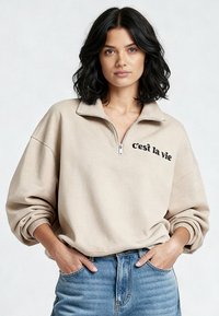 Maglione beige con zip a quarti e la scritta "c'est la vie" in nero, con vestibilità comoda e polsini a coste, abbinato a jeans azzurri chiaro.