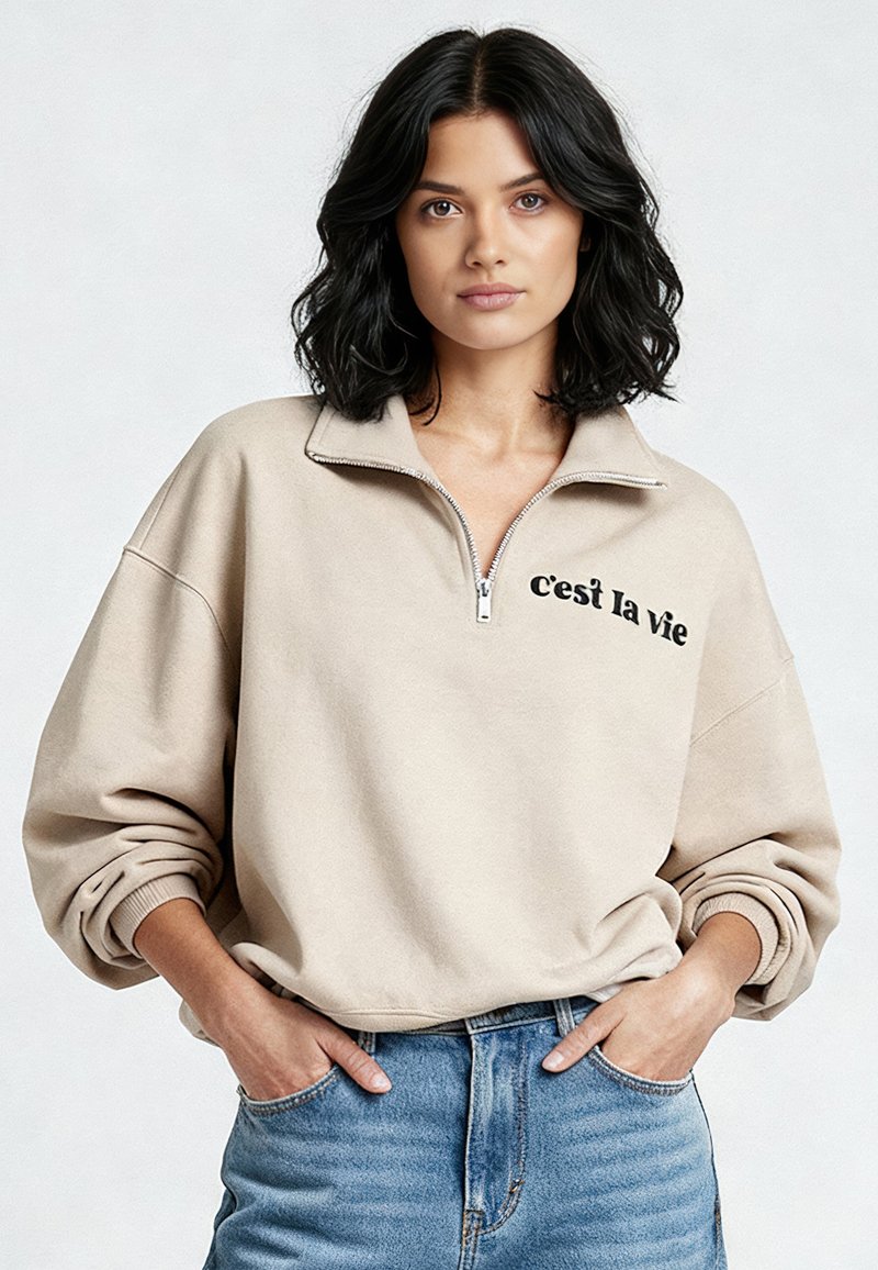 Maglione beige con zip a quarti e la scritta "c'est la vie" in nero, con vestibilità comoda e polsini a coste, abbinato a jeans azzurri chiaro.