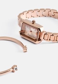 Relógio de pulso em rosa dourado com caixa octogonal, mostrador transparente e números romanos, acompanhado de uma pulseira combinando com um pequeno detalhe em gema.