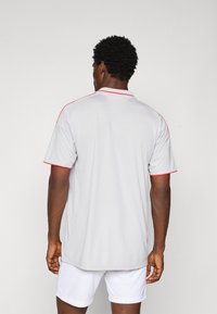 Un homme de dos portant un t-shirt de sport gris clair avec des accents rouges et un short de sport blanc, contre un fond clair.