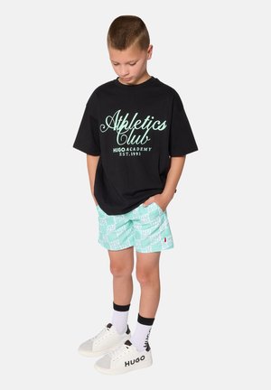 Schwarzes übergroßes T-Shirt mit grünem "Athletics Club"-Aufdruck, mintgrüne karierten Shorts, weiße Turnschuhe mit grauen Akzenten und schwarze Socken.