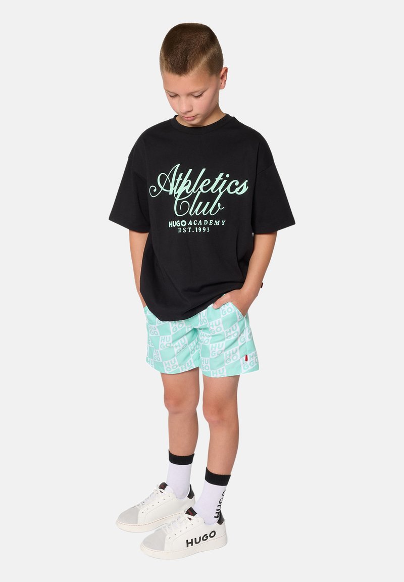 T-shirt noir oversize avec inscription verte "Athletics Club", short à carreaux menthe, baskets blanches avec accents gris et chaussettes noires.