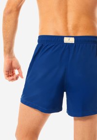 Pantaloni da bagno blu con vita elastica e etichetta in tessuto sul retro. Tessuto liscio, design fino al ginocchio, stile semplice senza fantasie.