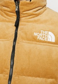 Brun manchesterjacka fram med svart dragkedja och vitt broderat The North Face-logga på bröstet.
