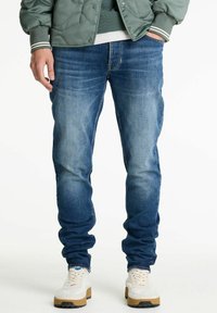 Jeans in denim blu con lavaggio sbiadito, fit slim, design a cinque tasche e dettagli cuciti sulle cuciture, abbinati a sneaker bianche.