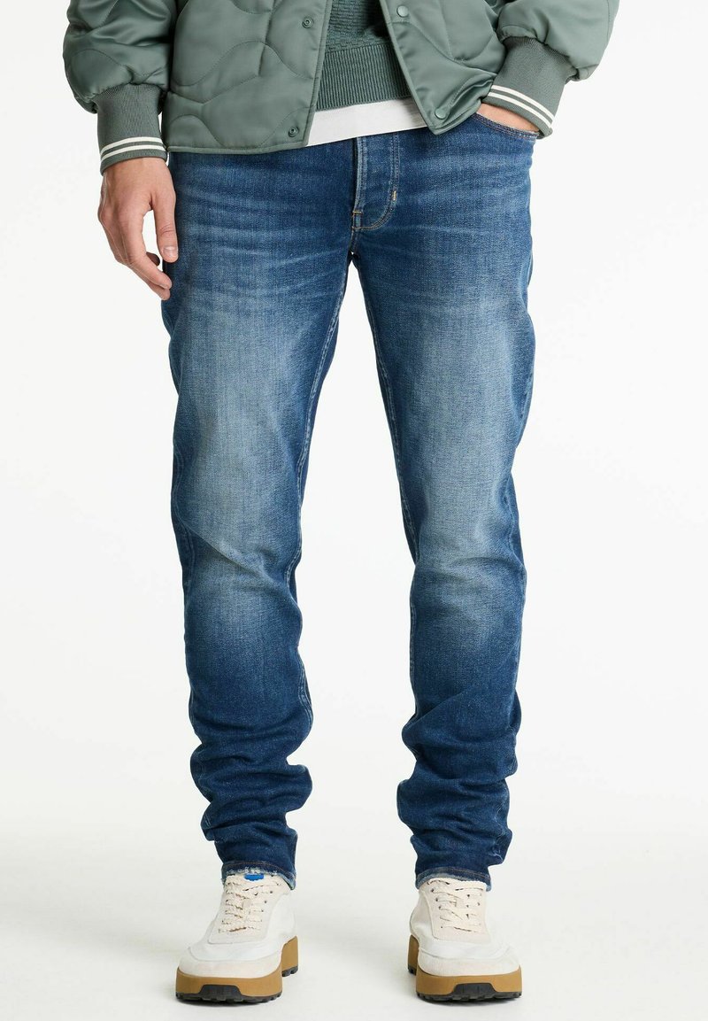 Jeans de mezclilla azul con un acabado desgastado, corte slim, diseño de cinco bolsillos y detalles cosidos en las costuras, combinados con zapatillas blancas.