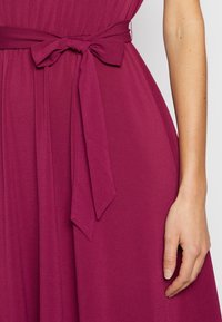 Magenta sukienka o gładkiej teksturze, z wysmuklonym talią i związanym paskiem, z płynącym materiałem, który elegancko się układa.