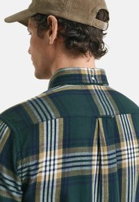 Man in een groen, blauw, beige en wit geruit shirt en een lichtbruine corduroy honkbalpet, gezien van achteren en de linkerkant.