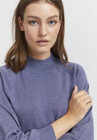 Violets adīts džemperis ar augstu apkakli, raglanu piedurknēm un ribotiem aproces. Mīksta tekstūra ar smalkām krāsas variācijām.