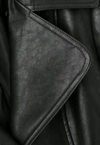 Dettaglio di una giacca di pelle nera con finitura testurizzata, rever affilati e cuciture prominenti lungo i bordi.