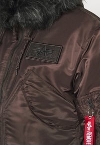 Hnědá bunda Alpha Industries se zipem vpředu, šikmou náprsní kapsou, černým koženým límcem a červenou visačkou "Odstraňte před letem" na zipu rukávu.