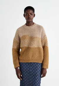 Stickad tröja i beige och senapsgul, med rund hals, lös passform och strukturerat mönster. Buren över en vadderad blå kjol.