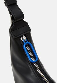 Musta nahkareppu, jossa on metallivetoketju ja sininen muovivetotappi. Hihna on säädettävä ja valmistettu vastaavasta nahasta.