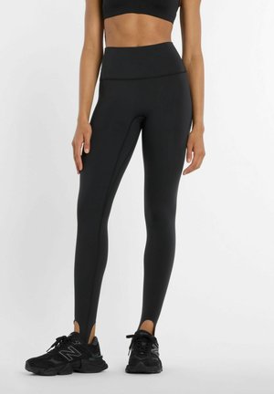 Donna che indossa leggings neri a vita alta con staffe e scarpe sportive nere, in piedi davanti a uno sfondo semplice.