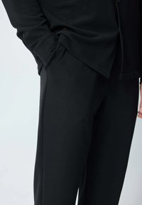 Haut noir à manches longues avec une coupe décontractée, texture côtelée et ourlet légèrement courbé, accompagné de pantalons noirs assortis ayant une finition lisse.