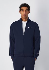 Giacca a zip blu navy con colletto alto, maniche lunghe e logo sul petto. Tessuto liscio, polsini a costine e apertura frontale con zip.