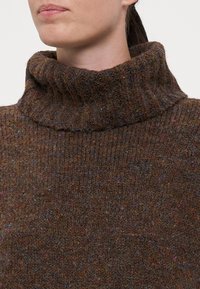 Brauner Wollrollkragenpullover mit einer groben Rippenstruktur und einem hohen, umgeschlagenen Kragen. Kleine bunte Fäden sind im gesamten Stoff sichtbar.