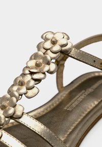 Μεταλλικά χρυσά σανδάλια με ανθισμένες διακοσμήσεις, με επίπεδα λουριά και λεία υφή. Η εσωτερική σόλα φέρει λεπτομέρεια με το σήμα της μάρκας.