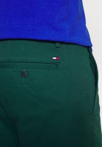 Shorts di cotone verdi con una tasca posteriore dotata di un bottone nero e un piccolo etichetta rossa, bianca e blu. La maglietta è blu.