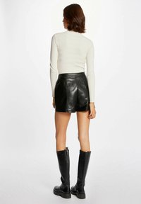Zwarte leren shorts met een hoge taille, gecombineerd met een fitted witte geribbelde lange mouwen top en kniehoge zwarte laarzen, gezien van achteren.