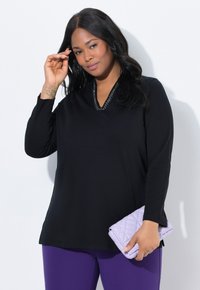 Vrouw met lang zwart haar, gekleed in een zwarte top met lange mouwen en paarse broek, houdt een gewatteerde lavendelkleurige clutch in één hand.