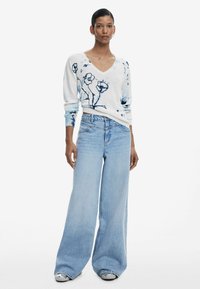 Weißer Pullover mit blauem Blumenmuster, V-Ausschnitt und langen Ärmeln, kombiniert mit hoch taillierten, weit geschnittenen hellblauen Jeans. Strukturiertes Material.