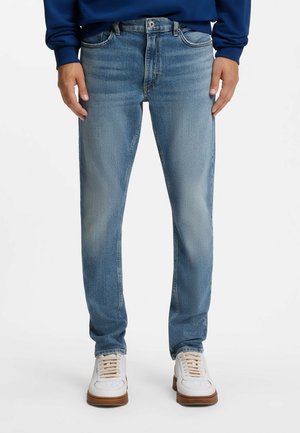 Mand iført lyseblå jeans med lige pasform, hvide sneakers med brune såler og en blå langærmet top, står mod en hvid baggrund.