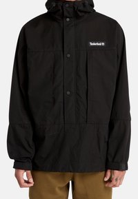 Svart, lätt, huva jacka med knappstängning, två framfickor och "Timberland"-logotyp. Har en avslappnad passform och långa ärmar.
