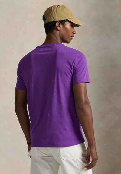 Μωβ βαμβακερό μπλουζάκι με κοντά μανίκια και λαιμόκοψη σε στρογγυλό σχήμα, φορεμένο με ανοιχτόχρωμο παντελόνι cargo. Η πίσω όψη δείχνει μια λεία υφή.