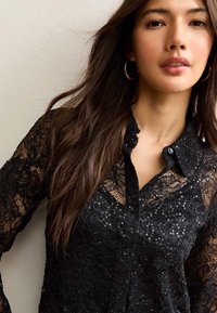 Czarna koronkowa bluzka z falbaniastym kołnierzykiem, z przodu zapinaną na guziki i akcentami z cekinów. Przezroczysta tekstura z długimi rękawami i misternymi wzorami.