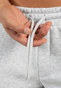 Lichtgrijze joggingbroek met een zachte textuur, voorzien van een trekkoord in de taille. Een hand houdt twee gevlochten trekkoorden bij de taille.