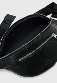 Zwarte leren heuptas met een getextureerd oppervlak, voorzien van een zilveren ritssluiting en een contrasterende zwarte riem met logodesign. De binnenkant is zwart gevoerd.