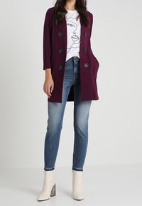 Bordeaux dubbelknäppt kappa med svarta knappar, bärs över en vit grafisk t-shirt och blå skinny jeans, kombinerat med vita klackade ankelstövlar.