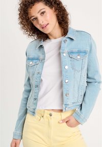 Chaqueta de mezclilla en azul claro con un diseño ajustado, botones de hardware plateado, dos bolsillos en el pecho y costuras en contraste, combinada con una camiseta blanca.