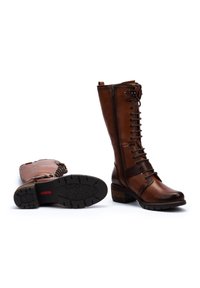 Bottes en cuir marron, hauteur genou, avec lacets, fermeture éclair latérale, détails de sangle et talon épais ; une botte est debout, l'autre est à plat sur sa semelle.